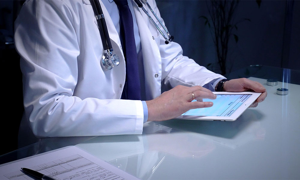 A physician wearing a stethoscope reviews medical information on a tablet beside paperwork.